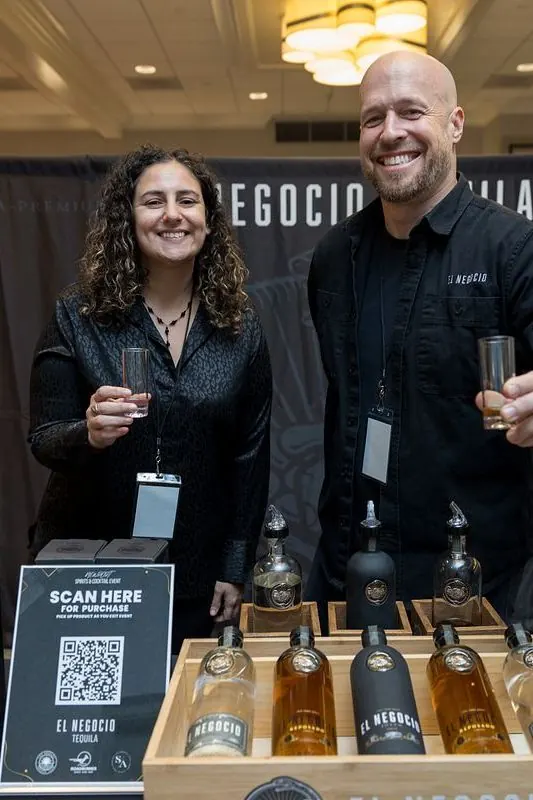 Two smiling exhibitors holding tasting glasses at the El Negocio Tequila booth, beside a “Scan here for purchase” sign and bottles of tequila