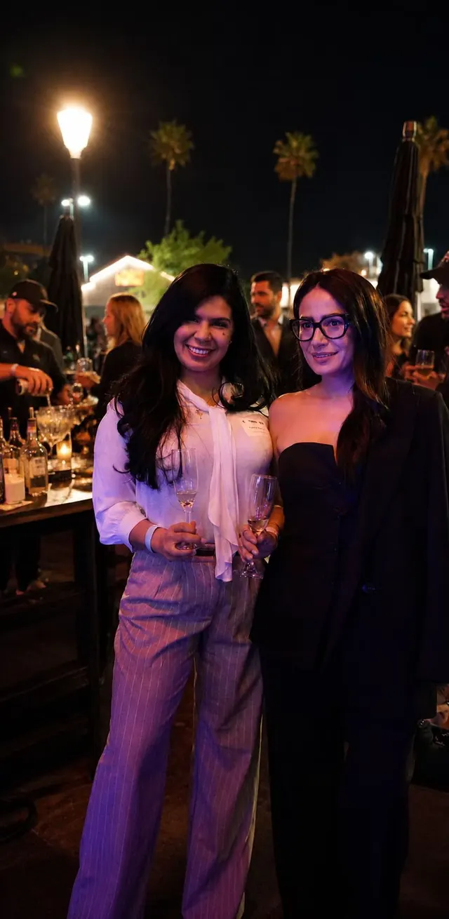 Two guests smiling with stemmed glasses at an outdoor evening reception with warm lights and palm trees
